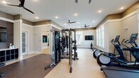 Located at 3000 Dominion Ridge Circle in Morrisville, NC, this single-floor fitness center is thoughtfully designed to support a full range of workout routines. The space is organized into distinct functional zones, creating a structured yet open environment for exercise.

The main workout area is generously sized and filled with natural light streaming through large windows fitted with horizontal blinds. Multiple ceiling fans throughout the space ensure comfortable air circulation during workouts. Recessed lighting provides consistent, even illumination across all areas.

The cardio zone lines the windowed perimeter and is equipped with a row of treadmills, elliptical trainers, and stair climbers, all oriented to take advantage of the natural light. A dedicated spin studio features several stationary cycling bikes and is enclosed with glass partitions and floor-to-ceiling mirrors, contributing to an open, airy feel.

The strength training area is anchored by a professional-grade cable machine system and a power rack with an attached bench, positioned on a dark rubberized floor mat. A mirrored wall and wall-mounted flat-screen TVs enhance the space throughout.

A wood-paneled accent wall adds a warm, contemporary design element, complemented by a built-in cubby storage unit. French doors provide access between sections of the facility, reinforcing the space's clean, upscale aesthetic.