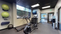 This fitness facility offers a focused and functional workout environment designed to meet essential exercise needs. The space features a dark, grid-style ceiling that lends a modern, industrial aesthetic, complemented by recessed panel lighting that ensures the area is well-lit throughout. Large windows along one wall flood the interior with natural light and provide views of the surrounding greenery, creating an energizing atmosphere for exercise. The flooring consists of durable rubber gym matting, practical for both cardio and strength training activities. Equipment includes SportsArt cardio machines — a treadmill and an elliptical trainer — positioned to take advantage of the natural light from the windows. A dumbbell rack with a selection of free weights is available for strength training, and a wall-mounted flat-screen television provides entertainment during workouts. A TRX suspension training system is anchored to the wall, adding versatility for bodyweight exercises. The space is accessed through a frosted glass door, which prominently displays the facility's logo, contributing to a polished, professional presentation. The overall design balances functionality with a clean, contemporary aesthetic, making it a practical and inviting space for daily fitness routines.