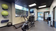 This fitness facility offers a focused and functional workout environment designed to meet essential exercise needs. The space features a dark, grid-style ceiling that lends a modern, industrial aesthetic, complemented by recessed panel lighting that ensures the area is well-lit throughout. Large windows along one wall flood the interior with natural light and provide views of the surrounding greenery, creating an energizing atmosphere for exercise. The flooring consists of durable rubber gym matting, practical for both cardio and strength training activities. Equipment includes SportsArt cardio machines — a treadmill and an elliptical trainer — positioned to take advantage of the natural light from the windows. A dumbbell rack with a selection of free weights is available for strength training, and a wall-mounted flat-screen television provides entertainment during workouts. A TRX suspension training system is anchored to the wall, adding versatility for bodyweight exercises. The space is accessed through a frosted glass door, which prominently displays the facility's logo, contributing to a polished, professional presentation. The overall design balances functionality with a clean, contemporary aesthetic, making it a practical and inviting space for daily fitness routines.