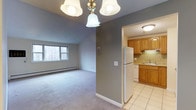 This single-floor unit offers a practical and comfortable layout designed to maximize its well-proportioned footprint. The living area features neutral gray walls, plush carpeting, and a classic chandelier overhead, creating a welcoming atmosphere. A window overlooking a wooded exterior view allows natural light to filter into the space, while a baseboard heating system ensures year-round comfort.

Adjacent to the living area, the kitchen is accessible through a wide doorway, providing an open and connected feel. The kitchen is outfitted with warm-toned wood cabinetry, granite countertops, and a decorative tile backsplash that adds a touch of character. White appliances, including a full-sized refrigerator and dishwasher, complement the space, while tile flooring adds durability and ease of maintenance. A flush-mount ceiling light brightens the area efficiently.

The overall design balances function and comfort, with the neutral color palette throughout creating a cohesive aesthetic. The unit is well-suited for those seeking a manageable, move-in-ready space with essential amenities in a convenient single-level configuration.