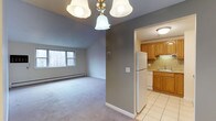 This single-floor unit offers a practical and comfortable layout designed to maximize its well-proportioned footprint. The living area features neutral gray walls, plush carpeting, and a classic chandelier overhead, creating a welcoming atmosphere. A window overlooking a wooded exterior view allows natural light to filter into the space, while a baseboard heating system ensures year-round comfort.

Adjacent to the living area, the kitchen is accessible through a wide doorway, providing an open and connected feel. The kitchen is outfitted with warm-toned wood cabinetry, granite countertops, and a decorative tile backsplash that adds a touch of character. White appliances, including a full-sized refrigerator and dishwasher, complement the space, while tile flooring adds durability and ease of maintenance. A flush-mount ceiling light brightens the area efficiently.

The overall design balances function and comfort, with the neutral color palette throughout creating a cohesive aesthetic. The unit is well-suited for those seeking a manageable, move-in-ready space with essential amenities in a convenient single-level configuration.