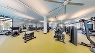 This expansive fitness center spans a generously sized single floor, providing ample room for a full complement of exercise equipment and dedicated training zones. The layout is thoughtfully organized, separating cardio and strength training areas to maximize usability and traffic flow.

The main workout floor features yellow rubber flooring, housing an extensive selection of strength machines, free weights, barbells, benches, and cable stations from brands including Hoist. A dedicated cardio zone lines the perimeter, equipped with treadmills, ellipticals, and stationary bikes positioned in front of large floor-to-ceiling windows that frame outdoor views and flood the space with natural light.

A separate functional training area with grey carpet flooring accommodates a power rack, sled, and open-floor exercises. Throughout the facility, bold typographic murals featuring fitness-themed words in blue, orange, and yellow energize the space visually. Large ceiling fans and linear LED lighting maintain comfort and visibility across all zones. Structural columns are integrated cleanly into the floor plan without disrupting movement. The overall design is modern, vibrant, and well-equipped for diverse fitness needs.