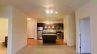 This single-floor residence offers a thoughtfully designed open-concept layout that seamlessly connects the kitchen and living areas. The kitchen is a standout feature, showcasing rich dark wood cabinetry that provides ample storage, complemented by sleek granite countertops and a full suite of stainless steel appliances, including a refrigerator, range, and microwave. A central kitchen island adds both prep space and a natural gathering point, while a row of elegant pendant lights hanging above creates a warm, inviting ambiance. Track lighting in the adjoining living area further enhances the brightness of the space. The transition from the tiled kitchen to the warm hardwood flooring, and into the carpeted living area, defines each zone while maintaining a cohesive, flowing aesthetic. Light, neutral wall tones throughout keep the interior feeling open and airy. The layout is practical and efficient, with clearly defined entryway access and interior doors leading to additional rooms. Overall, this space combines contemporary finishes with functional design, making it well-suited for everyday living.