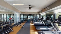 This contemporary fitness center is thoughtfully designed to provide a comprehensive workout environment within a single, open-plan floor. The space features a dual-zone flooring layout, combining gray carpet with cork-toned rubber flooring that visually separates the cardio area from the strength and free-weight zones. Ceiling fans and recessed linear lighting maintain a comfortable, well-lit atmosphere throughout.

The cardio section is lined with a full row of treadmills, StairMaster steppers, and stationary bikes, all positioned to take advantage of the floor-to-ceiling glass windows and doors that line the perimeter, flooding the space with natural light and offering views of the surrounding outdoor environment. A dual set of glass entry doors connects the gym directly to the exterior.

The strength training area is stocked with a complete rack of dumbbells, adjustable benches, medicine balls, and a cable crossover machine, catering to a wide range of free-weight and functional training exercises. Full-length mirrors are mounted along the walls, enhancing the sense of space while allowing users to monitor their form. The overall ambiance is clean, modern, and purposefully functional, making this fitness center a well-rounded facility suited for all levels of fitness training.
