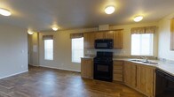 This single-story home showcases an open-concept layout that seamlessly connects the kitchen and living areas, creating a spacious and functional environment. The kitchen is equipped with medium-toned wood cabinetry featuring upper and lower storage, granite-look countertops, and a full suite of black appliances, including a gas range, over-the-range microwave, and a sink positioned along the perimeter. The cabinetry extends to provide ample storage, complemented by a clean, neutral color palette on the walls. Wood-style hard flooring runs throughout the visible areas, adding warmth and durability to the space. Multiple windows dressed with decorative valances allow natural light to filter into both the kitchen and the adjacent living area, keeping the space bright and inviting. Flush-mount ceiling light fixtures provide consistent overhead illumination throughout. The open flow between the kitchen and living space makes the layout well-suited for everyday living and entertaining. The overall design reflects a practical and comfortable interior with a cohesive, neutral aesthetic throughout.