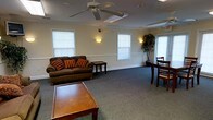This single-floor space is designed as a welcoming common area, offering a well-organized and functional layout suited for lounging and social interaction. The room features neutral-toned walls accented by a white chair rail that runs along the perimeter, giving the space a clean, structured appearance. Soft gray carpeting covers the floor throughout, adding warmth and comfort underfoot.

A ceiling fan with a light fixture is centrally positioned, providing both air circulation and ambient lighting. Wall-mounted sconces on either side of the room cast a warm, inviting glow, complementing the natural light that filters in through multiple windows fitted with white horizontal blinds. The windows are evenly distributed along the walls, contributing to a bright and airy atmosphere.

The seating area includes a dark brown sofa adorned with decorative orange and red pillows, paired with a wooden end table. A rectangular wooden coffee table anchors the foreground, while a dining-style table and chairs occupy one side of the room, providing additional utility. A decorative potted tree adds a natural element near the entrance, enhancing the overall warmth and livability of the space.