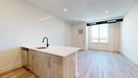 Located at 55 Washington Street in East Orange, NJ, this thoughtfully designed one-bedroom, one-bathroom residence offers a clean, contemporary aesthetic throughout. The open-plan kitchen and living area serves as the heart of the unit, anchored by a stylish kitchen island with light wood cabinetry, a white quartz countertop, and a matte black gooseneck faucet. Stainless steel appliances, including a range and refrigerator, complement the modern design. Recessed lighting and a sleek linear pendant fixture illuminate the space, while light wood-tone flooring extends seamlessly throughout, adding warmth and continuity. The living area benefits from a large window that invites an abundance of natural light and frames open scenic views of the surrounding landscape. The spacious bedroom features floor-to-ceiling windows on multiple walls, flooding the room with daylight and offering expansive outdoor views. Crisp white walls and light flooring maintain the unit's bright, airy character. The bathroom features terrazzo-style flooring, providing a refined contrast to the rest of the unit's design. A dedicated closet and in-unit laundry add practical convenience. The overall ambiance is fresh, modern, and move-in ready.