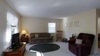 This single-floor residence offers a comfortable and classically styled living environment. The living room serves as the central gathering space, featuring light-toned walls and a neutral carpet that create a warm, inviting atmosphere. Natural light filters through a curtained window, brightening the room and highlighting the space's clean, simple lines. The room is furnished with a patterned upholstered sofa, a dark upholstered armchair, and a traditional wooden rocking chair, lending the space a relaxed, homey character. A round dark area rug anchors the seating arrangement and adds visual contrast to the light flooring. Wooden side tables and a table lamp provide functional accent pieces, while a small collection of framed artwork adorns the walls, contributing to the room's understated, classic aesthetic. The layout is open and uncluttered, allowing for easy movement throughout the space. The smooth white ceiling and softly lit interior further enhance the sense of ease and comfort. Overall, this residence presents a practical and welcoming single-level layout well-suited to everyday living.