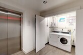 Located at 1326 Florida Ave NE in Washington, DC, this compact shared utility space serves as both a functional laundry room and connecting hallway for building residents. The laundry area is equipped with commercial-grade Speed Queen washers and dryers, complemented by a wall-mounted water heater and clearly posted usage instructions — offering a convenient, coin-free laundry experience. The walls are finished in clean white paint with concrete block construction, maintaining a practical and easy-to-maintain environment. The hallway area connects key circulation points within the building and features wood-look luxury vinyl plank flooring that lends a contemporary touch throughout. A stainless steel elevator with modern paneling provides accessible vertical transit, while a bold, graphic black-and-white patterned accent wall adds a striking decorative element to the corridor. Sliding and hinged dark-paneled doors contribute to the modern aesthetic, and a security intercom panel is conveniently positioned near the entry. The space is efficiently laid out, maximizing functionality within its compact footprint while maintaining a clean, well-finished appearance consistent with a modern residential building.
