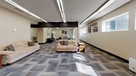 Located at 1001 E 12th St in Gillette, Wyoming, this single-floor facility offers a well-organized and functional layout designed for communal use. The expansive main lounge area is anchored by patterned carpet tile flooring and illuminated by fluorescent linear lighting, with a bold dark structural beam adding an architectural accent overhead. Multiple seating arrangements with sofas, armchairs, and end tables create comfortable gathering zones throughout the open floor plan. A pool table is visible in the background, adding a recreational element to the space. Colorful artwork adorns the neutral-toned walls, contributing a welcoming atmosphere. A dedicated dining nook features a classic round wooden table with Windsor-style chairs, positioned beneath a row of horizontal windows that bring in natural light and views of the surrounding neighborhood. The kitchen is fully equipped with dark wood cabinetry, white appliances including a refrigerator, electric range, and double sink, set over durable tile flooring — well-suited for group meal preparation. Additional corridors lead to private rooms or offices, and the overall layout supports a variety of community-oriented activities. The textured ceilings and consistent neutral wall tones throughout give the space a clean, organized, and practical character.
