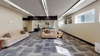 Located at 1001 E 12th St in Gillette, Wyoming, this single-floor facility offers a well-organized and functional layout designed for communal use. The expansive main lounge area is anchored by patterned carpet tile flooring and illuminated by fluorescent linear lighting, with a bold dark structural beam adding an architectural accent overhead. Multiple seating arrangements with sofas, armchairs, and end tables create comfortable gathering zones throughout the open floor plan. A pool table is visible in the background, adding a recreational element to the space. Colorful artwork adorns the neutral-toned walls, contributing a welcoming atmosphere. A dedicated dining nook features a classic round wooden table with Windsor-style chairs, positioned beneath a row of horizontal windows that bring in natural light and views of the surrounding neighborhood. The kitchen is fully equipped with dark wood cabinetry, white appliances including a refrigerator, electric range, and double sink, set over durable tile flooring — well-suited for group meal preparation. Additional corridors lead to private rooms or offices, and the overall layout supports a variety of community-oriented activities. The textured ceilings and consistent neutral wall tones throughout give the space a clean, organized, and practical character.
