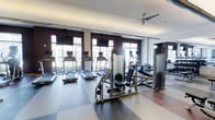 The fitness center at Woodmont Metro at Metuchen Station is a thoughtfully designed, single-level amenity space that offers residents a well-equipped environment for their workout routines. The room features large windows along one wall, flooding the space with natural light and providing views of the surrounding exterior, creating an open and energizing atmosphere.

The facility is outfitted with a comprehensive selection of modern exercise equipment, including multiple treadmills equipped with individual display screens, ideal for cardio training. A multi-station cable and weight machine anchors the center of the room, offering versatile strength-training options, while additional resistance and weight equipment lines the perimeter. Mounted flat-screen televisions are positioned above the windows, allowing exercisers to stay entertained during their workouts.

The flooring features a multi-toned tile pattern that defines the space with a clean, contemporary aesthetic. Overhead, a ceiling fan provides additional air circulation, complementing the room's bright and comfortable environment. The dark accent walls provide a modern contrast to the white ceiling, lending the space a polished, upscale feel well-suited to a fitness-focused community.
