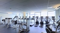 This contemporary fitness center is designed to deliver a premium workout experience in a bright, open environment. The space is lined with expansive floor-to-ceiling glass windows along one full wall, flooding the interior with natural light and offering views of the surrounding cityscape — creating an energizing atmosphere for exercise.

The cardio section is generously stocked with a row of treadmills and elliptical cross-trainers, each unit equipped with individual digital display screens, allowing users to track performance metrics or enjoy entertainment during their workout. Strength and resistance training is supported by a selection of professional-grade cable and weight machines positioned throughout the floor, providing options for a full-body training routine.

The flooring consists of dark, durable fitness-grade material suited for high-traffic athletic use, while the suspended tile ceiling with integrated lighting complements the natural light from the windows. The overall layout is thoughtfully organized, providing ample space between equipment to ensure comfortable and safe use. The clean, modern aesthetic and well-maintained equipment reflect a facility built for serious fitness use, making it suitable for a wide range of users, from casual exercisers to dedicated athletes.