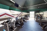 This fitness center is a well-equipped, single-level workout space designed to support a range of cardio-focused exercise routines. The room's most visually striking feature is its bold geometric wall mural, rendered in deep red, grey, white, and black chevron and arrow patterns that wrap around the space, creating an energetic and motivating atmosphere. Full-length mirrors line portions of the walls, visually expanding the room and allowing users to monitor their form during workouts.

The facility houses a variety of cardio machines, including multiple treadmills, upright and recumbent stationary bikes, elliptical trainers, and a rowing machine, all arranged efficiently on dark rubber flooring that provides both durability and traction. Overhead, a distinctive green-paneled angled ceiling features multiple ceiling fans to promote air circulation, while a wall-mounted air conditioning unit offers additional climate control. Mounted flat-screen televisions are positioned for easy viewing during exercise sessions.

A gated opening at one end of the room introduces natural light and provides a glimpse of an outdoor pool area beyond, offering a refreshing connection to the exterior environment and enhancing the overall workout experience.