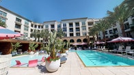 This expansive outdoor amenity space is designed to deliver a resort-style experience within a modern residential complex. At its heart is a generously sized swimming pool with crystal-clear turquoise water, framed by wide sandstone-toned tile decking that provides ample room for lounging. Rows of white chaise lounges line the pool's perimeter, shaded by distinctive pink-and-white striped umbrellas and pergola structures that lend a vibrant, upscale atmosphere.

Surrounding the pool, tall palm trees and bold tropical plantings — including large banana-leaf plants in oversized white planters accented with red florals — create a lush, resort-like ambiance. Manicured artificial turf zones offer additional relaxation areas, including a sunken lounge with built-in wood-paneled bench seating, colorful cushions, and a stone fire pit, ideal for evening gatherings.

Private wood-paneled cabanas provide a more intimate retreat, each furnished with comfortable seating, a flat-screen television, ceiling fan, and chevron-patterned rugs. Hedged greenery walls offer natural privacy screening between areas. The entire space is framed by an elegant multi-story building featuring arched architectural details, completing the polished, resort-inspired aesthetic.
