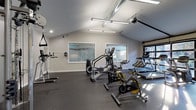 This fitness center is a comprehensively outfitted exercise facility designed to support a full range of workout routines. The space features vaulted ceilings with exposed ductwork that lend it an open, industrial character, while large garage-style glass doors along one wall flood the interior with natural light and provide views of the surrounding outdoor environment. Linear pendant lighting supplements the natural light, ensuring the space remains bright and well-illuminated throughout.

The rubber-floored main area is organized to accommodate multiple training disciplines. A dedicated cardio zone includes treadmills, elliptical trainers, stationary bikes, and cycling bikes, all arranged to take advantage of the natural light from the glass doors. A strength training section features a multi-functional cable machine, a dumbbell rack with a full range of weights, and additional resistance equipment. A functional training and stretching area includes exercise mats, stability balls in multiple sizes, and medicine ball storage.

Mirrors are strategically placed along the walls to assist with form and to enhance the sense of space. A wall-mounted water fountain provides convenient hydration access. Informational signage is posted near the entrance, offering fitness guidelines for users. The overall design is clean and modern, with neutral gray walls and durable flooring that create a focused, professional atmosphere suited for effective training sessions.