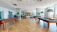 This contemporary amenity lounge is designed to offer a dynamic and multifunctional social environment across a single, generously proportioned floor. Striking herringbone-patterned hardwood flooring flows throughout the space, unifying its distinct zones while adding warmth against the sleek, modern palette of exposed concrete columns, white tile ceilings, and neutral walls accented by colorful artwork.

The main open area serves as a game and recreation hub, featuring a shuffleboard table, a billiards table, and a foosball table, all suspended beneath pendant lighting. Floor-to-ceiling windows frame sweeping panoramic city views, bathing the space in abundant natural light.

Adjacent lounge areas offer varied seating arrangements, including a large sectional sofa positioned directly against the expansive windows for unobstructed skyline views, along with accent chairs and casual tables. A dedicated media lounge features a built-in wood-paneled entertainment wall with a flat-screen TV, plush blue and yellow accent chairs, and a patterned area rug.

A lively dining and social nook displays vibrant blue seating around round white tables beneath colorful cone pendant lights, complemented by playful wall murals. A bar-height kitchen counter area with stools provides a casual gathering point. Together, these zones create a well-appointed, versatile lounge suited for both relaxed socializing and active recreation.