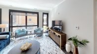 This thoughtfully designed single-floor residence offers a refined blend of contemporary style and functional living. The open living area is anchored by large, black-framed windows that flood the space with natural light and frame views of the surrounding urban environment. A herringbone-patterned area rug, teal accent chairs, and a warm walnut media console with brass accents lend the room a sophisticated yet inviting character.

The kitchen is outfitted with high-gloss white cabinetry, granite countertops, stainless steel appliances including a gas range, and a peninsula with seating — ideal for casual dining or entertaining. Dark hardwood flooring adds warmth and continuity throughout the main living spaces.

Two well-appointed bedrooms offer comfortable retreats, each featuring plush carpeting, dark-framed windows, and ample storage through sleek, dark-toned dressers. The rooms are styled with coordinating textiles and accent artwork, contributing to the space's cohesive modern aesthetic.

The bathroom is a standout feature, showcasing floor-to-ceiling marble-style tile in the walk-in glass shower, hexagonal floor tile, a dark vanity with granite countertop, and an illuminated mirror — delivering a spa-like experience. Crown molding and a neutral grey palette throughout tie the entire residence together with elegance and precision.