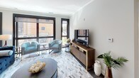 This thoughtfully designed single-floor residence offers a refined blend of contemporary style and functional living. The open living area is anchored by large, black-framed windows that flood the space with natural light and frame views of the surrounding urban environment. A herringbone-patterned area rug, teal accent chairs, and a warm walnut media console with brass accents lend the room a sophisticated yet inviting character.

The kitchen is outfitted with high-gloss white cabinetry, granite countertops, stainless steel appliances including a gas range, and a peninsula with seating — ideal for casual dining or entertaining. Dark hardwood flooring adds warmth and continuity throughout the main living spaces.

Two well-appointed bedrooms offer comfortable retreats, each featuring plush carpeting, dark-framed windows, and ample storage through sleek, dark-toned dressers. The rooms are styled with coordinating textiles and accent artwork, contributing to the space's cohesive modern aesthetic.

The bathroom is a standout feature, showcasing floor-to-ceiling marble-style tile in the walk-in glass shower, hexagonal floor tile, a dark vanity with granite countertop, and an illuminated mirror — delivering a spa-like experience. Crown molding and a neutral grey palette throughout tie the entire residence together with elegance and precision.