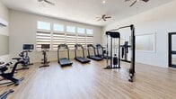 This fitness center offers a generously sized, open workout space designed to accommodate a diverse range of exercise equipment and training styles. The interior is finished with light hardwood-style flooring that spans the entire area, contributing to a clean, modern aesthetic throughout.

The main workout floor is organized to support both cardio and strength training. A row of treadmills lines one wall beneath a bank of clerestory-style windows fitted with layered roller shades, allowing natural light to filter in while maintaining privacy. Alongside the treadmills, stationary cycling bikes with individual screen displays are arranged near a wall-mounted flat-screen TV and a split-system air conditioning unit. A dual cable resistance machine is centrally positioned, providing a functional strength training station. Elliptical trainers round out the cardio offerings on the opposite side of the room.

Multiple ceiling fans are distributed across the ceiling, ensuring consistent air circulation. Recessed lighting supplements the natural light from the windows. Interior glass panel windows connect to an adjacent room, adding visual openness. The overall design is bright, neutral, and purposefully laid out to maximize usable training space while maintaining a comfortable and functional environment.
