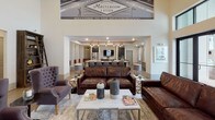 The Ironwood Masteroom & Kitchen is a thoughtfully designed single-floor communal gathering space that blends comfort, style, and functionality. The main lounge area commands immediate attention with its soaring ceilings, dramatic double-height windows, and an impressive tiered chandelier that serves as a central focal point. Rich hardwood-style flooring runs throughout, anchoring the warm, welcoming aesthetic.

The seating area is furnished with deep leather sofas, tufted accent chairs, and marble-top coffee tables, creating an inviting atmosphere for both relaxed socializing and casual gatherings. A classic fireplace with a white mantel adds an elegant touch to one wall, while a mounted flat-screen TV provides entertainment options. Clerestory windows positioned high along the walls flood the space with abundant natural light.

Adjacent to the lounge, the kitchen and bar area features a long island with counter seating, pendant lighting, and stainless steel appliances, making it well-suited for communal dining or events. A separate, more intimate lounge nook is also present, offering a quieter setting with its own seating arrangement and tray ceiling detail.

Large French doors along one wall open directly to an outdoor pool area, seamlessly connecting the interior with the exterior. The overall design balances a neighborhood tavern aesthetic with modern residential comfort, making it an inviting hub for community life.
