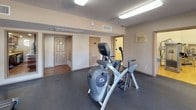 Located at 4701 American Blvd in Fort Worth, TX, this fitness and recreation facility offers a comprehensive workout environment designed to support a wide range of exercise needs. The space is organized across a single floor and includes several distinct functional zones.

The main cardio area is lined with large windows that allow natural light to fill the room and provide views of the outdoor surroundings. This section is outfitted with Cybex upright bikes, arc trainers, and treadmills, all positioned along the windowed wall for an open, airy feel. Durable rubber flooring throughout this zone provides comfort and safety underfoot.

Adjacent to the cardio area, a spacious strength training section features a comprehensive lineup of Cybex selectorized machines targeting major muscle groups, including leg press, chest press, and cable systems. This area transitions into a racquetball court, distinguished by its classic hardwood flooring with red boundary markings and tall, smooth white paneled walls that reach impressive heights, creating a bright and clean playing environment.

A dedicated stretching and floor exercise area completes the facility, finished with dark rubber flooring and floor-to-ceiling mirrors that enhance the sense of space. The facility is well-lit throughout with overhead fluorescent lighting, and multiple access points ensure convenient circulation between zones.