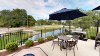 This single-floor space is highlighted by a standout outdoor patio area that seamlessly blends comfort and scenic appeal. The patio is finished with a solid concrete surface and furnished with decorative cast iron outdoor seating, including a round table with chairs and a navy blue market umbrella, providing a comfortable spot for dining or relaxing outdoors. A gas grill is conveniently positioned nearby, making the space well-suited for outdoor entertaining. A decorative fire feature is also present, adding ambiance to the outdoor setting. The patio is bordered by a classic black wrought iron fence that encloses the space while maintaining open sightlines to the serene waterway just beyond. Neatly trimmed round shrubs and manicured landscaping line the fence, adding a polished, well-maintained aesthetic. The waterfront backdrop offers peaceful views of a calm pond or canal, with mature trees and green space visible in the distance under an open sky. The overall character of the space is inviting and functional, with a strong emphasis on outdoor living and the natural surroundings. The combination of thoughtful landscaping, quality outdoor furnishings, and direct water views creates a tranquil and practical environment ideal for leisure and gatherings.