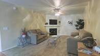 This single-story space centers around a well-appointed living room designed for comfort and functionality. The room features a corner fireplace with a clean white mantel, serving as a natural focal point and providing both warmth and visual interest. A wall-mounted flat-screen television is positioned above the mantel, creating a practical media and gathering area. The living room is finished with plush wall-to-wall carpeting in a neutral beige tone, contributing to a warm and inviting atmosphere. A ceiling fan with an integrated light fixture is centrally mounted, offering both air circulation and ambient lighting. The seating arrangement includes two sofas and an additional chair, complemented by glass-top accent tables with decorative metalwork bases. The walls feature a subtle textured finish with soft, sweeping tonal variations that add depth and character to the space. Decorative touches, including potted plants and framed wall art, enhance the livability of the room. The overall aesthetic is cohesive and neutral, making the space adaptable to a variety of furnishing styles.