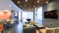 This thoughtfully designed single-floor lounge space makes an immediate impression with its distinctive slatted wood ceiling, where warm-toned wooden slats are interspersed with recessed lighting to create a visually dynamic overhead feature. The ceiling design adds depth and architectural interest while casting a warm, ambient glow throughout the interior.

The layout is organized into distinct seating areas, encouraging both social interaction and relaxed lounging. A comfortable sectional sofa anchors the central gathering zone, complemented by upholstered accent chairs and wooden coffee tables that add a natural warmth to the contemporary palette. A linear fireplace is built into a sleek accent wall, providing a cozy focal point and adding to the inviting atmosphere.

Toward the perimeter, floor-to-ceiling windows allow abundant natural light to pour in, offering views of the surrounding urban environment. Concrete columns punctuate the space while reinforcing its modern aesthetic. A wall-mounted flat-screen television and a dining or meeting table visible in the background suggest the space serves multiple functions. The overall ambiance is polished yet welcoming, blending modern design with comfortable, functional furnishings.
