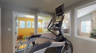 The fitness center is a thoughtfully designed single-level space outfitted with professional-grade Matrix cardio equipment, including an elliptical trainer and treadmill, positioned to make the most of the available floor area. Dark, durable flooring throughout the workout space provides a sturdy and practical surface suited for active use. The neutral gray walls give the room a clean, focused atmosphere, while white trim detailing adds a polished contrast.

A standout architectural feature is the series of interior windows that connect the fitness area to neighboring spaces. One set of windows overlooks a warmly lit lounge featuring vaulted ceilings, a stone fireplace, and comfortable seating—offering an inviting visual backdrop during workouts. Another interior window and open doorway face a vibrant children's play area, distinguished by bold yellow walls, colorful patterned rugs, and bright natural light, adding an energetic and community-friendly feel to the overall environment.

Practical amenities include a paper towel dispenser, waste bin, a wall-mounted mirror, and a fire extinguisher, ensuring both convenience and safety. A thermostat provides climate control, and a glass-paneled entry door connects the space to the broader facility. The layout and design reflect a functional, community-centered fitness environment.