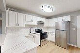 This single-floor, 2-bedroom, 1-bathroom home offers a thoughtfully designed layout with a cohesive aesthetic of cool gray walls, crisp white trim, and light hardwood flooring across the main living areas. The open living room connects seamlessly to the kitchen via a pass-through window, creating a sense of openness and easy interaction between spaces. The kitchen is a standout feature, equipped with stainless steel appliances including a refrigerator, electric range, and dishwasher, complemented by white shaker cabinetry with matte black hardware, a subway tile backsplash, and marble-look countertops — all tied together with durable hardwood-style flooring. The primary bedroom is a comfortable, well-sized room featuring soft carpet, a ceiling fan with light fixture, and a window that allows natural light to filter in. The second bedroom, also carpeted, offers a quieter, cozy atmosphere. The bathroom is finished in a clean, all-white palette with a tiled tub-shower combination, a vanity with a white countertop, and a window for natural ventilation. The hallway entry area features hardwood-style flooring and a front door with sidelight window, providing a welcoming transition into the home.