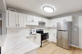 This single-floor, 2-bedroom, 1-bathroom home offers a thoughtfully designed layout with a cohesive aesthetic of cool gray walls, crisp white trim, and light hardwood flooring across the main living areas. The open living room connects seamlessly to the kitchen via a pass-through window, creating a sense of openness and easy interaction between spaces. The kitchen is a standout feature, equipped with stainless steel appliances including a refrigerator, electric range, and dishwasher, complemented by white shaker cabinetry with matte black hardware, a subway tile backsplash, and marble-look countertops — all tied together with durable hardwood-style flooring. The primary bedroom is a comfortable, well-sized room featuring soft carpet, a ceiling fan with light fixture, and a window that allows natural light to filter in. The second bedroom, also carpeted, offers a quieter, cozy atmosphere. The bathroom is finished in a clean, all-white palette with a tiled tub-shower combination, a vanity with a white countertop, and a window for natural ventilation. The hallway entry area features hardwood-style flooring and a front door with sidelight window, providing a welcoming transition into the home.