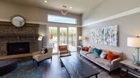 This single-floor common area is designed with a contemporary aesthetic, combining open-plan living with thoughtfully curated zones for gathering, dining, and relaxing. The space is unified by rich hardwood-style flooring, elevated ceilings with crown molding details, and a neutral gray-toned color palette that lends a clean, modern character throughout.

The lounge area anchors one side of the space, featuring a plush velvet sofa accented with bold teal and rust throw pillows, complementary seating chairs, and a rustic wood coffee table. A stone fireplace adds warmth and texture, while large-scale abstract artwork contributes a vibrant focal point. Clerestory windows positioned above the patio doors draw in natural light without sacrificing wall space.

The dining area accommodates multiple glass-top round tables with dark upholstered seating, and a wall-mounted flat-screen television adds functionality. French doors open directly to an outdoor patio equipped with additional seating, a grill station, and an umbrella-shaded table.

The kitchen is outfitted with dark espresso cabinetry, granite countertops, a full-length blue tile backsplash, stainless steel appliances, a built-in beverage refrigerator, and a breakfast bar with leather stools beneath statement pendant lighting. Together, these elements create a cohesive and inviting communal environment.