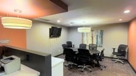 This single-floor professional space is designed to facilitate productive meetings and collaborative work. The conference area serves as the centerpiece of the space, featuring a round conference table surrounded by ergonomic mesh-back office chairs, providing comfortable seating for group discussions and presentations. A wall-mounted flat-screen television is positioned for easy viewing by all participants, making it well-suited for presentations and video conferencing needs. Overhead, a prominent drum-style pendant light fixture provides warm, focused illumination over the meeting table, complementing the recessed ceiling lights that ensure the room is evenly and efficiently lit. The neutral-toned walls create a clean, professional atmosphere, accented by a colorful abstract artwork that adds a touch of creativity to the environment. A partial partition wall with a dark wood accent band adds architectural interest and subtly defines the space. Patterned carpeted flooring grounds the room with texture and warmth. The overall ambiance strikes a balance between functionality and professionalism, making this space a practical and inviting setting for business meetings, team collaboration, or client consultations.