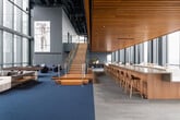 This sophisticated two-floor amenity space occupies a prominent position high within a glass-curtain tower, defined throughout by floor-to-ceiling windows that frame sweeping panoramic views of the city skyline and waterfront beyond. Blue carpeting flows consistently across both levels, anchoring a cohesive design language that blends warm wood tones with cool, muted grays and blues.

The main lounge area features soaring double-height ceilings with a slatted wood ceiling detail that adds warmth and acoustic character. Generous seating arrangements — including large sectional sofas in soft blues and whites — are positioned to take full advantage of the dramatic views, while curated artwork and pendant lighting contribute a refined, gallery-like atmosphere.

A dedicated conference room offers a substantial live-edge wood table surrounded by upholstered swivel chairs, pendant lighting, and a wall-mounted display, all set against a backdrop of expansive city views. A recreation area includes a full-size billiards table alongside lounge seating and a large display screen. Additional areas feature casual bar-height seating with a high counter, flexible lounge nooks with modular furniture, and a wide perimeter corridor lined with individual chairs positioned directly before the panoramic windows — ideal for quiet contemplation or informal gatherings.