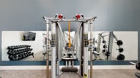 This expansive fitness center is thoughtfully designed to accommodate a wide range of workout routines and fitness levels. The space features soaring double-height ceilings throughout much of the facility, with large clerestory windows that flood the interior with natural light and create an open, energizing atmosphere. Neutral-toned carpet tiles cover the floors, complemented by crisp white walls accented with deep teal-blue paneling that adds a modern, polished aesthetic.

The main workout floor is outfitted with professional-grade Hoist strength training machines, including a functional trainer cable system, as well as a selection of cardio equipment such as treadmills and ellipticals. A dedicated free weights area features a full dumbbell rack and benches, alongside a wall-mounted flat-screen TV for entertainment during workouts. An open functional training zone offers a wall-mounted rig with suspension trainers, battle ropes, and a medicine ball rack, positioned beneath dramatic floor-to-ceiling windows.

A separate spin studio houses multiple cycling bikes and its own wall-mounted display screen. A mezzanine-level area, accessed by a modern staircase with sleek metal railings, provides additional cardio equipment and overlooks the main floor below. Full-length mirrors throughout enhance the sense of space and support proper form during training.