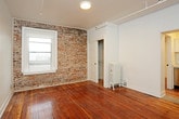 This single-floor space blends classic architectural character with modern updates throughout. The main living area is generously proportioned, showcasing rich, warm hardwood floors that run throughout the entire unit. A standout exposed brick wall anchors the living space, adding texture and an industrial charm, while a window allows natural light to fill the room. A cast-iron radiator provides a nod to the building's history.

The kitchen is efficiently laid out with clean white cabinetry, light quartz countertops, and stainless steel appliances including a gas range, over-the-range microwave, and full-size refrigerator. The design maximizes functionality within the compact footprint.

The bedroom continues the hardwood flooring and features its own cast-iron radiator, a sliding panel door, and a window with views of the surrounding neighborhood, providing natural light and ventilation.

The bathroom is finished in a crisp, all-white palette, featuring large-format vertically stacked subway tiles surrounding the soaking tub and shower combination. A wall-mounted sink with chrome fixtures and a medicine cabinet complete the clean, modern aesthetic. The hardwood flooring extends into the bathroom, connecting the space cohesively to the rest of the unit.