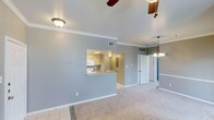 This single-floor residence offers a clean, open layout designed for comfortable everyday living. The main living area is finished with wall-to-wall carpeting and cool gray-toned walls, creating a calm and neutral ambiance throughout. Crown molding runs along the ceilings, adding a touch of classic architectural detail to the space. A ceiling fan provides practical comfort overhead, while a decorative chandelier near the dining area adds a subtle elegant touch.

The kitchen is thoughtfully connected to the living area via a pass-through opening with a raised breakfast bar counter, allowing for easy interaction between spaces while maintaining a defined cooking zone. The kitchen features light wood cabinetry, white appliances, and its own ceiling fan, contributing to an efficient and functional layout. Tile flooring in the kitchen provides a durable, easy-to-clean surface that transitions smoothly into the carpeted living and dining areas.

Ample natural light filters through the space, complemented by recessed vents and flush-mount lighting that ensure year-round climate control and visibility. The entryway area is visible from the living room, offering a clear and accessible flow through the home. Overall, this residence presents a well-maintained, move-in-ready interior with a cohesive design and practical single-level living.