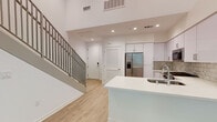 This contemporary two-story residence is designed with a clean, modern aesthetic defined by bright white walls, light wood-look flooring, and recessed lighting throughout. The main level opens into an airy living and entry area distinguished by soaring double-height ceilings and large windows that allow abundant natural light to fill the space. A sleek staircase with gray metal balusters connects the two floors seamlessly.

The kitchen is thoughtfully designed with white shaker-style cabinetry reaching to the upper walls, a subway tile backsplash, and a suite of stainless steel appliances including a gas range, microwave, and a French door refrigerator with an in-door water and ice dispenser. A large white island with an integrated sink serves as both a functional workspace and a visual centerpiece of the main living area.

The residence includes two bathrooms, each finished to a high standard. One features a glass-enclosed walk-in shower with white subway tile, built-in open shelving, a white quartz vanity, and an LED-backlit mirror. The second bathroom includes accessibility-friendly grab bars, a tall storage cabinet, a matching quartz countertop vanity, and a second LED-backlit mirror. Additional rooms on the upper level offer bright, versatile spaces with windows framing outdoor greenery, completing this well-appointed and functional home.