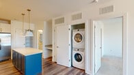 Located at 1000 Lakeside Terrace Ct in Wake Forest, NC, this well-designed one-bedroom, one-bathroom unit offers a thoughtful layout with contemporary finishes throughout. The open-concept kitchen and living area serves as the heart of the home, showcasing white shaker cabinetry, a white subway tile backsplash, stainless steel appliances, and a striking blue kitchen island topped with a light countertop, illuminated by stylish gold pendant lights. Wood-look flooring flows throughout the main living areas, adding warmth and cohesion. A dedicated nook provides built-in desk space, ideal for a home office setup. A conveniently tucked-away closet houses a stacked washer and dryer, offering in-unit laundry. The living area opens directly onto a private balcony through a glass-panel door, bringing in abundant natural light and outdoor views of the surrounding landscape. The spacious bedroom features plush carpet, a ceiling fan, wide windows with outdoor views, and direct access to the full bathroom. The bathroom is elegantly appointed with a white vanity, gold hardware, a large framed mirror, and a walk-in glass-enclosed shower with floor-to-ceiling tile surround. A walk-in closet with wire shelving completes the bedroom suite, providing generous storage.