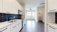 Located at 222 West Blvd in Charlotte's vibrant 28203 corridor, this single-floor residence showcases a clean, modern design with high-quality finishes at every turn. The open-concept kitchen is a standout feature, outfitted with crisp white cabinetry, sleek black hardware, light stone countertops, and a striking herringbone-patterned tile backsplash in deep navy. Stainless steel appliances, including a range, over-the-range microwave, and refrigerator, complete the well-appointed culinary space. Rich wood-look flooring runs throughout the unit, anchoring the contemporary aesthetic. The living area is bathed in natural light from large windows that frame views of the surrounding Charlotte skyline and greenery, complemented by a ceiling fan overhead. A spacious walk-in closet with built-in shelving provides generous storage. The bathroom is thoughtfully designed with a full soaking tub, hexagonal mosaic tile surround, matte black fixtures, a white vanity with stone countertop, and wood-tone flooring. Additional utility is offered through a dedicated electrical panel area with washer/dryer hookups. The overall ambiance is bright, airy, and modern, making this Charlotte residence a polished urban retreat.