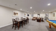 Located at 2-48 Grove Ave in Cedar Grove, NJ, this single-floor community living room is a well-appointed recreational lounge designed for socializing and entertainment. The space is finished with neutral-toned walls, clean white baseboards, and wall-to-wall patterned carpeting that adds texture underfoot while maintaining a polished aesthetic throughout.

The room is divided into distinct activity zones. A dining area anchored by a dark wood table and matching cross-back chairs provides seating for group gatherings, while a wall-mounted flat-screen TV nearby adds a media element to the space. The lounge area features plush, oversized brown leather sofas arranged around a wooden coffee table, creating a comfortable spot for relaxation and conversation.

A full-size billiard table with a bold blue felt surface and ornate wooden legs serves as the room's centerpiece, offering a classic recreational focal point. Adjacent to it, a cue rack keeps equipment neatly organized. A compact wet bar with white cabinetry and dark countertops is tucked into one corner, providing a convenient refreshment station.

Recessed ceiling lighting illuminates the entire space evenly, while a curated collection of framed classic film posters — including Jaws, Reservoir Dogs, The Professional, GoodFellas, and Rocky — lends the room a cinematic, pop-culture character that ties the entertainment theme together cohesively.