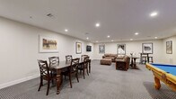 Located at 2-48 Grove Ave in Cedar Grove, NJ, this single-floor community living room is a well-appointed recreational lounge designed for socializing and entertainment. The space is finished with neutral-toned walls, clean white baseboards, and wall-to-wall patterned carpeting that adds texture underfoot while maintaining a polished aesthetic throughout.

The room is divided into distinct activity zones. A dining area anchored by a dark wood table and matching cross-back chairs provides seating for group gatherings, while a wall-mounted flat-screen TV nearby adds a media element to the space. The lounge area features plush, oversized brown leather sofas arranged around a wooden coffee table, creating a comfortable spot for relaxation and conversation.

A full-size billiard table with a bold blue felt surface and ornate wooden legs serves as the room's centerpiece, offering a classic recreational focal point. Adjacent to it, a cue rack keeps equipment neatly organized. A compact wet bar with white cabinetry and dark countertops is tucked into one corner, providing a convenient refreshment station.

Recessed ceiling lighting illuminates the entire space evenly, while a curated collection of framed classic film posters — including Jaws, Reservoir Dogs, The Professional, GoodFellas, and Rocky — lends the room a cinematic, pop-culture character that ties the entertainment theme together cohesively.