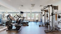 Located at 502 S Main St in Warrensburg, Missouri, this fitness center offers a comprehensive exercise facility designed to accommodate a full range of workout routines. The single-room layout is efficiently organized, maximizing available floor space while maintaining a clean, open feel throughout.

The room features durable rubber flooring ideal for both cardio and strength training activities. Natural light floods the space through a row of large windows along one wall, offering views of an outdoor pool area and greenery beyond. A ceiling fan provides air circulation, complementing the recessed lighting and ceiling-mounted HVAC vents that keep the environment comfortable during workouts.

Cardio equipment includes multiple treadmills from SOLE and Spirit, as well as an elliptical and a recumbent bike. For strength training, the facility is equipped with a multi-function cable machine, a squat rack with a bench press station, a full rack of dumbbells, and additional free weight plates. Wall-mounted mirrors on one side visually expand the space and assist with form during exercises. A wall-mounted television provides entertainment during workouts.

The room also includes a sanitizing station and posted fitness center rules near the bold blue entry door, reflecting an emphasis on cleanliness and community standards. Overall, this fitness center provides a practical and well-appointed environment for a variety of fitness goals.