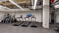 This fitness center offers a comprehensive workout environment spread across a single expansive floor. The space features an industrial aesthetic, with exposed ductwork, wood-beam ceilings, and bright linear LED lighting that illuminates the facility throughout. The flooring consists of durable dark rubber tiles, providing traction and comfort underfoot across all exercise zones.

The cardio area is well-appointed with multiple treadmills, stair climbers, and elliptical machines, complemented by wall-mounted flat-screen televisions for entertainment during workouts. A dedicated spin room houses Peloton bikes, each equipped with interactive screens, alongside an additional wall-mounted display.

The free weights section includes an extensive range of dumbbells neatly arranged on multi-tier racks, an adjustable workout bench, and kettlebells available in varying weights. Floor-to-ceiling mirrors line the walls in this area, allowing users to monitor their form. The open layout also accommodates cable and resistance machines, offering a full range of strength training options.

A water fountain is conveniently accessible within the main workout area. The white-painted walls contrast with the raw industrial ceiling elements, creating a clean and functional environment. The overall layout is open and spacious, allowing for comfortable movement between equipment zones.