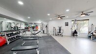 This fitness center offers a generously sized, single-floor workout space designed to accommodate a full range of exercise needs. The interior is finished with clean white walls and light-colored flooring, creating a bright and energizing atmosphere enhanced by recessed lighting and multiple ceiling fans that ensure comfortable airflow throughout. Large windows along one wall invite natural light and provide views of the surrounding exterior, adding an open, airy feel to the space.

The facility is thoughtfully divided into distinct workout zones. A dedicated cardio area houses several treadmills, elliptical trainers, and a stationary bike, all positioned in front of windows to make workouts more engaging. The strength training section features a comprehensive selection of cable and resistance machines suited for full-body conditioning. A free weights zone is stocked with a complete dumbbell rack, kettlebells, and medicine balls, accompanied by adjustable benches and wall-mounted mirrors that visually expand the space and assist with form.

A padded mat area with interlocking floor tiles provides a designated zone for stretching, functional training, or floor-based exercises. A striking, colorful geometric wall mural serves as a bold focal point near the entrance, adding a vibrant and motivating aesthetic touch. The overall ambiance is clean, modern, and functional — well-suited for a focused and effective workout experience.