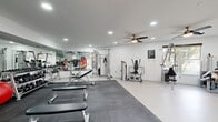 This fitness center offers a generously sized, single-floor workout space designed to accommodate a full range of exercise needs. The interior is finished with clean white walls and light-colored flooring, creating a bright and energizing atmosphere enhanced by recessed lighting and multiple ceiling fans that ensure comfortable airflow throughout. Large windows along one wall invite natural light and provide views of the surrounding exterior, adding an open, airy feel to the space.

The facility is thoughtfully divided into distinct workout zones. A dedicated cardio area houses several treadmills, elliptical trainers, and a stationary bike, all positioned in front of windows to make workouts more engaging. The strength training section features a comprehensive selection of cable and resistance machines suited for full-body conditioning. A free weights zone is stocked with a complete dumbbell rack, kettlebells, and medicine balls, accompanied by adjustable benches and wall-mounted mirrors that visually expand the space and assist with form.

A padded mat area with interlocking floor tiles provides a designated zone for stretching, functional training, or floor-based exercises. A striking, colorful geometric wall mural serves as a bold focal point near the entrance, adding a vibrant and motivating aesthetic touch. The overall ambiance is clean, modern, and functional — well-suited for a focused and effective workout experience.