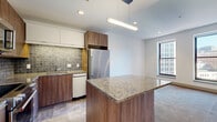 This well-appointed two-bedroom, two-bathroom residence offers a thoughtfully designed layout across a single floor. The open-concept kitchen anchors the living area, featuring warm wood cabinetry, a textured tile backsplash, stainless steel appliances including a full-size refrigerator, and a generous granite-topped island with pendant lighting above. The adjacent living area transitions to soft carpeting and is brightened by multiple windows framing elevated urban cityscape views.

The spacious primary bedroom is carpeted and receives abundant natural light through a pair of large windows overlooking the city skyline. The second bedroom is similarly finished with carpet flooring and neutral walls. Both rooms offer a clean, versatile canvas.

The primary bathroom is a standout feature, showcasing floor-to-ceiling stone-look tile, a frameless glass walk-in shower with a built-in bench and mosaic tile accent wall, and a floating vanity with a granite countertop and backlit mirror. The second bathroom is finished with coordinating stone tile and a wall-mounted toilet behind a sliding door.

A dedicated closet with built-in shelving and hanging rods doubles as a laundry area, housing a stacked washer and dryer unit. Recessed lighting and tile flooring complete the practical, polished interior throughout.