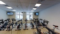 Located at 515 West Ave in Norwalk, Connecticut, this dedicated spin studio is a purpose-built fitness space designed to deliver an energizing and focused cycling experience. The room is outfitted with a full array of stationary spin bikes arranged in rows, providing ample capacity for group cycling classes. Full-length mirrors line the walls, visually expanding the space and allowing participants to monitor their form throughout their workout. Large windows along one wall usher in generous natural light, creating a bright and motivating atmosphere while offering views of the surrounding streetscape. The light wood-toned flooring complements the clean, neutral wall tones, lending the studio a fresh and modern feel. Overhead, a drop ceiling with recessed panel lighting ensures consistent, even illumination throughout the room. A wall-mounted flat-screen television is positioned at the front of the studio, ideal for displaying instructional content or class metrics. The space is equipped with a sprinkler system and ventilation to maintain comfort during high-intensity sessions. Overall, the studio strikes a balance between functional fitness design and an inviting, well-lit environment suited for both individual training and structured group classes.