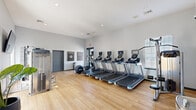Located at 7801 Scenic Hwy in Baton Rouge, Louisiana, this fitness center is a thoughtfully designed exercise facility occupying a single open floor. The space features light hardwood-style flooring throughout, complemented by neutral gray walls and high vaulted ceilings that create an airy, spacious atmosphere conducive to exercise.

The cardio section is prominently equipped with a row of modern treadmills featuring individual digital displays, alongside an elliptical trainer, providing ample options for aerobic workouts. On the strength training side, the facility includes a Precor multi-function cable and weight machine, an adjustable workout bench, a dumbbell rack, a stability ball, and a cable pull-down station, offering a comprehensive range of resistance training options.

Natural light flows generously into the space through large windows fitted with blinds, offering views of the surrounding outdoor environment. Recessed lighting supplements the natural light, ensuring the space remains well-lit throughout the day. Motivational wall art adds an energizing aesthetic touch to the environment.

Additional amenities include a water fountain, a vending machine, and clear access through glass-paneled doors. The overall layout is open and functional, allowing for comfortable movement between equipment zones, making this fitness center a practical and welcoming space for a full-body workout routine.